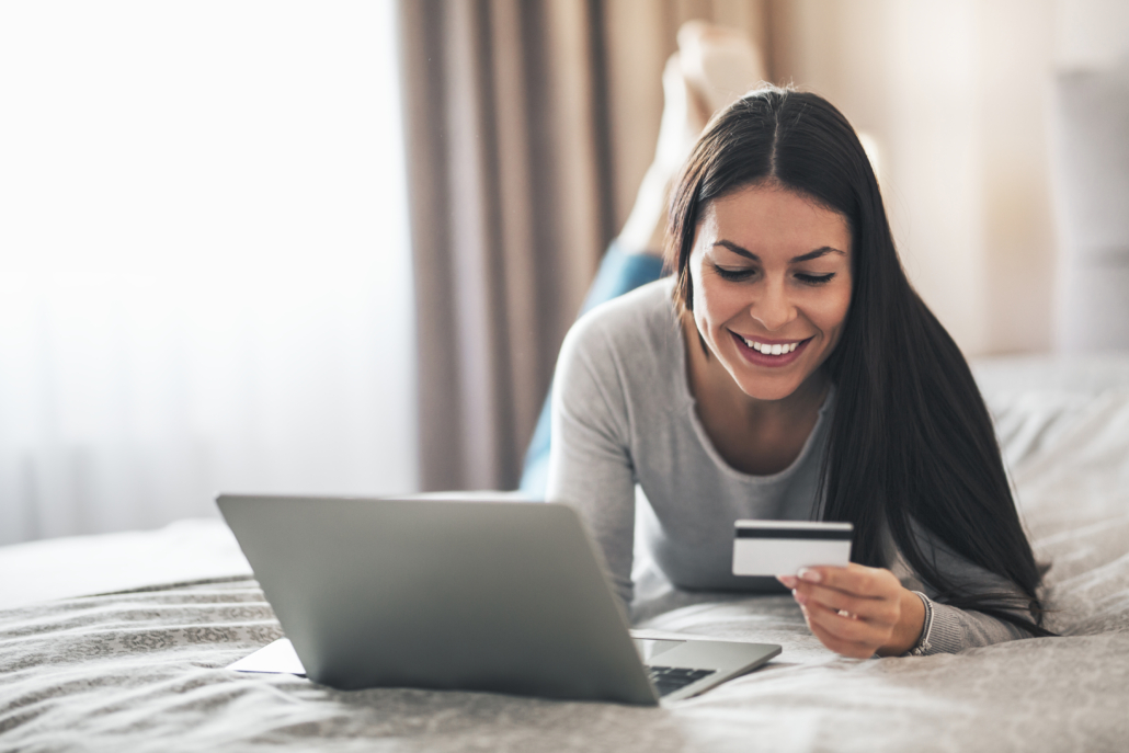 Beautiful girl lying on the bed and holding credit card while using laptop for online shopping.